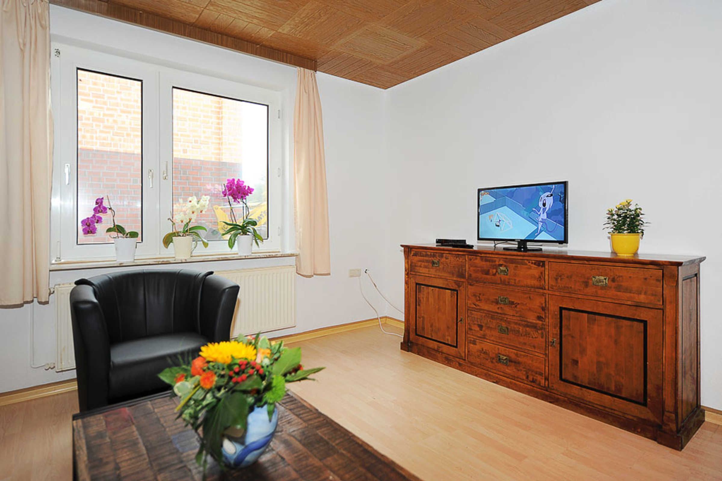 Un salón con un sofá de cuero negro, un armario de madera, un televisor y una ventana con flores.