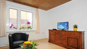 A living room with a black leather sofa, a wooden cabinet, a television, and a window with flowers. - Steering Wheel 2 - Apartments House Steering Wheel (Esens)