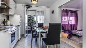 Une cuisine compacte dotée d’appareils électroménagers modernes, un coin repas avec une table en verre et des chaises noires, et un balcon agrémenté de rideaux violets.