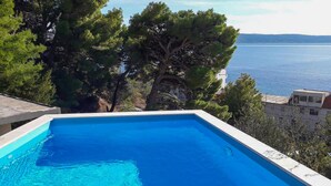 Une piscine rectangulaire à l’eau d’un bleu limpide, entourée d’arbres et de bâtiments, et offrant une vue sur une étendue d’eau.