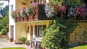 A two-story building with a balcony adorned with colorful flowers, a small outdoor seating area with a table and chairs, and a stone wall partially covered by ivy.