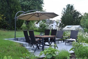 Une terrasse extérieure avec une table et des chaises, un grand parasol et, au fond, une serre.