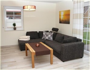 A modern living room with a grey sofa, a wooden coffee table, a hanging lamp, a painting, and a window with blinds.