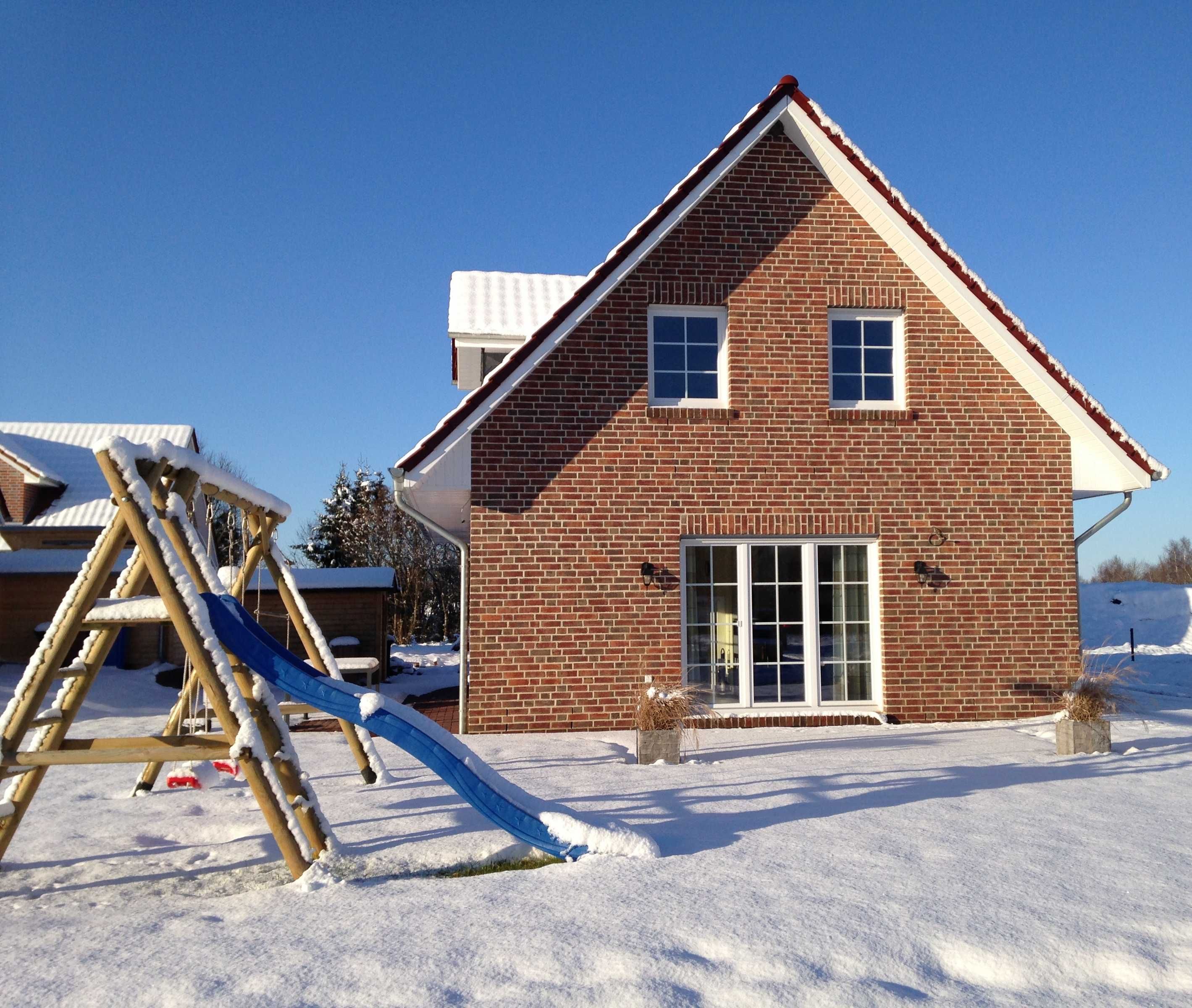 A brick house with a large window and a door, a wooden swing set, and snow on the ground.