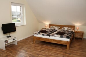 A bedroom with a wooden bed, a TV on a stand, a window, and a nightstand with a lamp. - Holiday house beach pearl (Garrel)