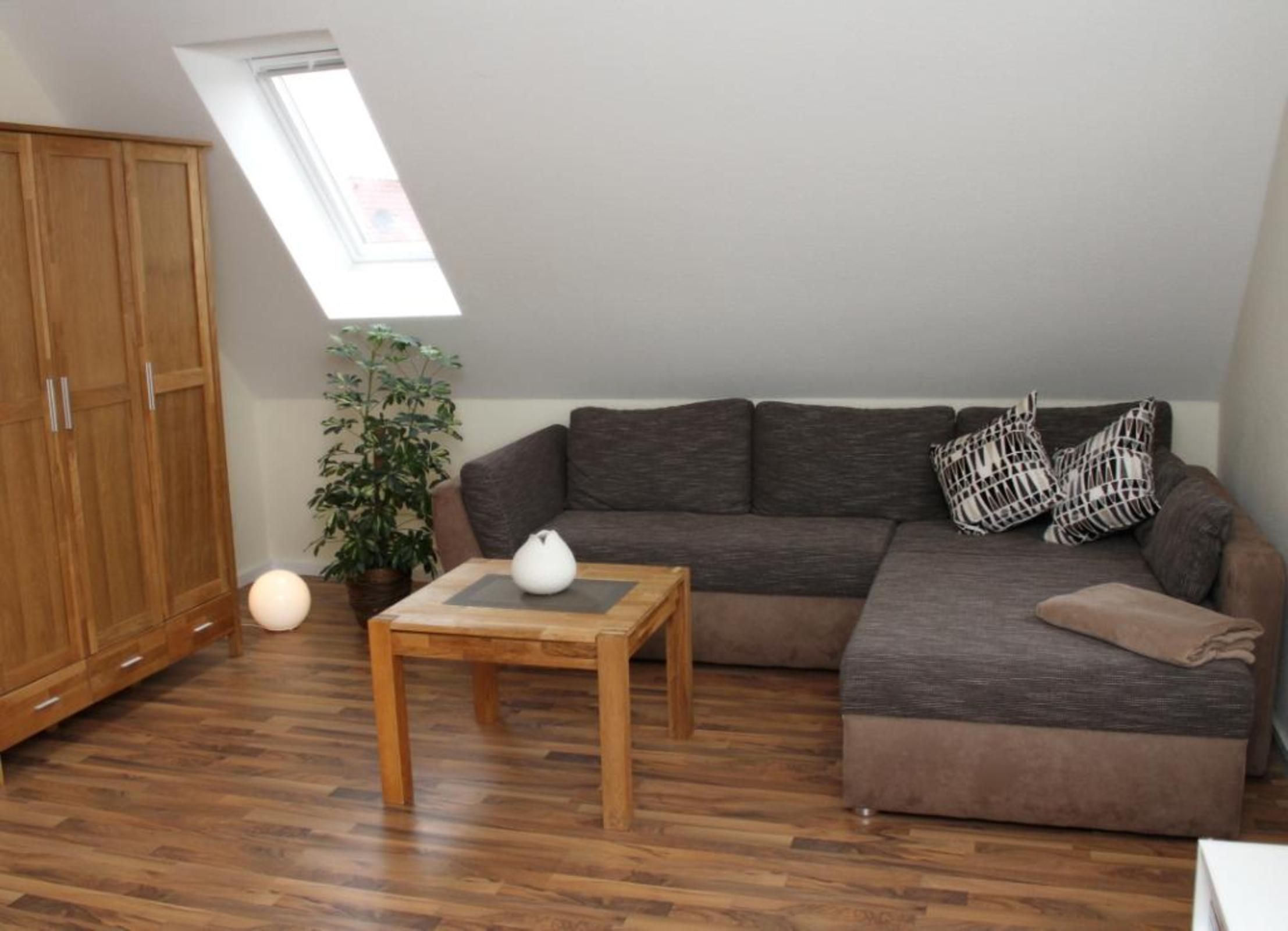 Quarto com piso de madeira, um sofá marrom, uma mesa pequena com um vaso branco, uma planta em vaso e um guarda-roupa grande.