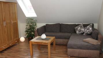 Quarto com piso de madeira, um sofá marrom, uma mesa pequena com um vaso branco, uma planta em vaso e um guarda-roupa grande.