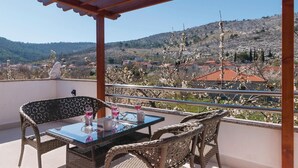 Un balcón con muebles de mimbre, una mesa de vidrio y vistas a un pueblo y colinas.