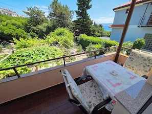 A balcony with a table and chairs, overlooking a garden and a building with a red roof.
