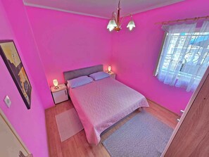Et rosa-farvet soveværelse med seng, sengeborde, lamper, et maleri og et vindue med gardiner.