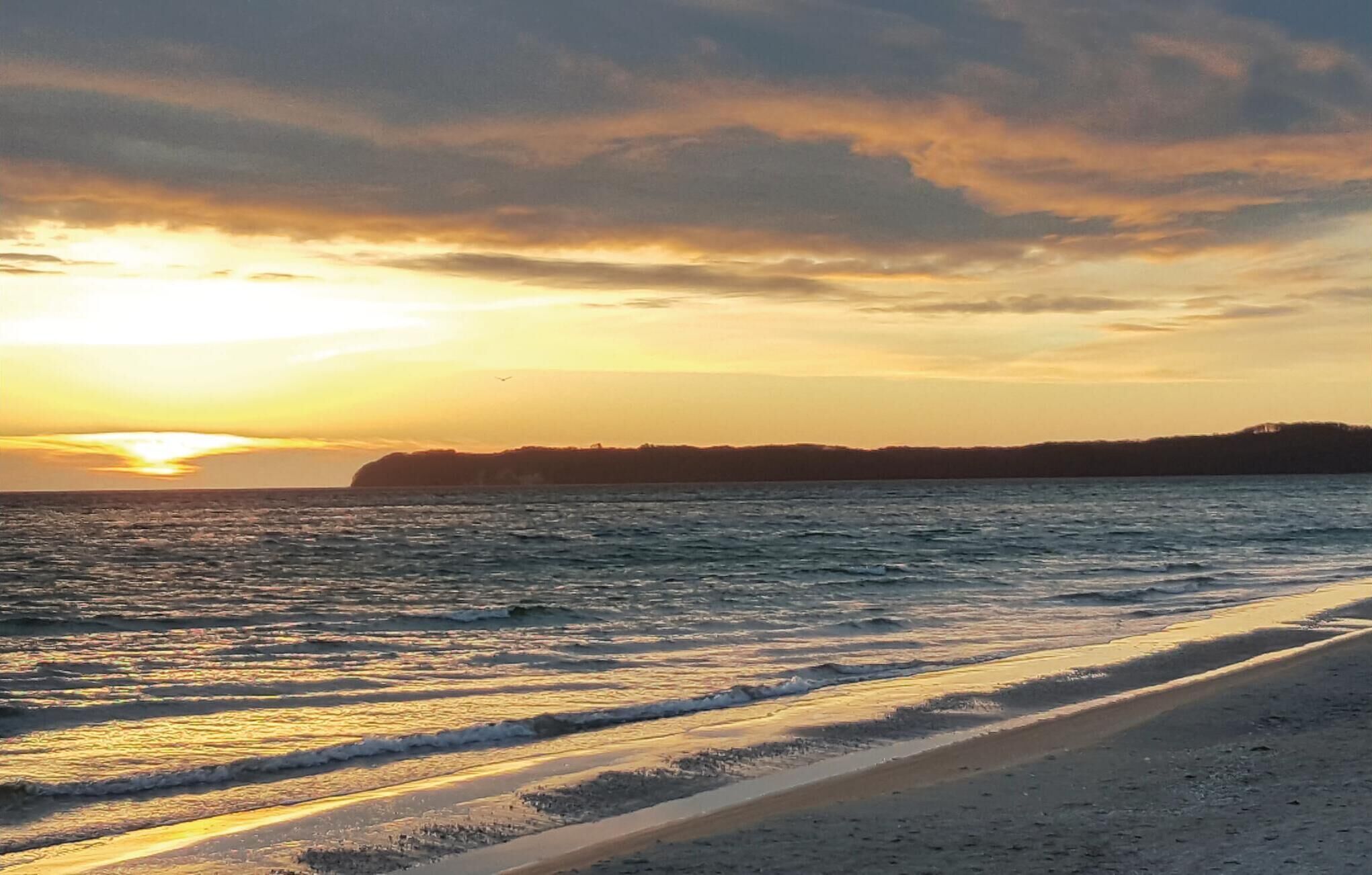 Une plage avec des vagues au coucher du soleil, une côte lointaine et un ciel dégagé.