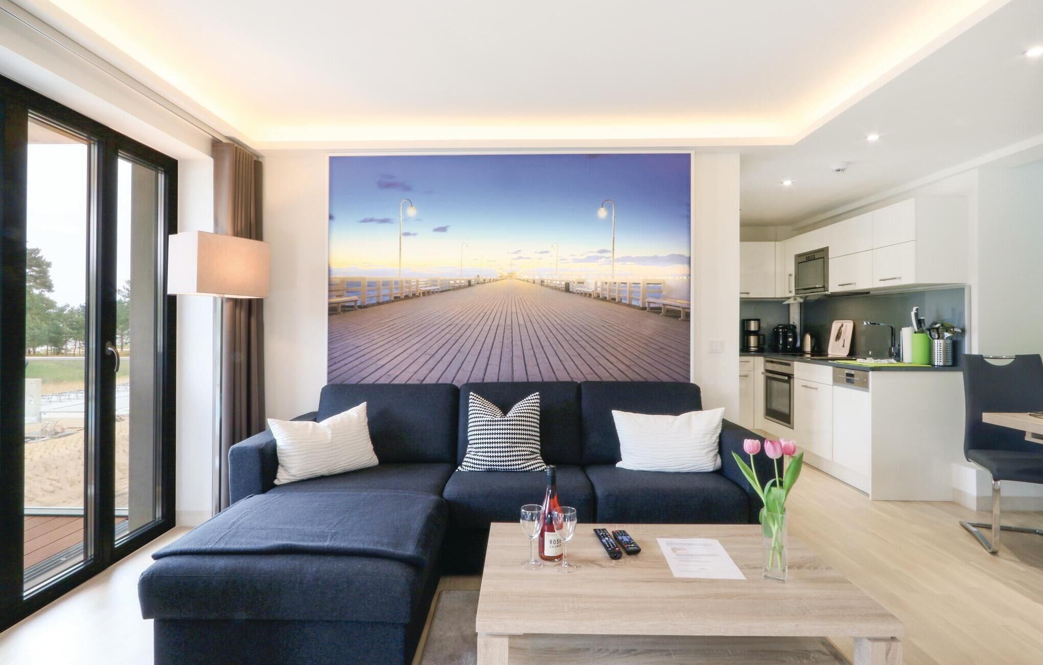 A modern hotel room with a large window, a sofa with cushions, a coffee table with a bottle of wine, glasses, and a remote control, and a kitchen area with a microwave and coffee maker.