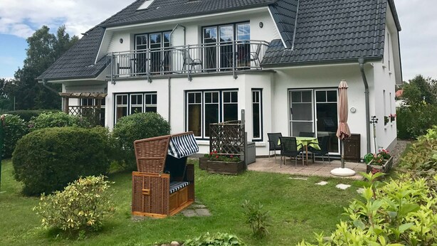 A two-story house with a balcony, large windows, and a garden area with outdoor furniture.