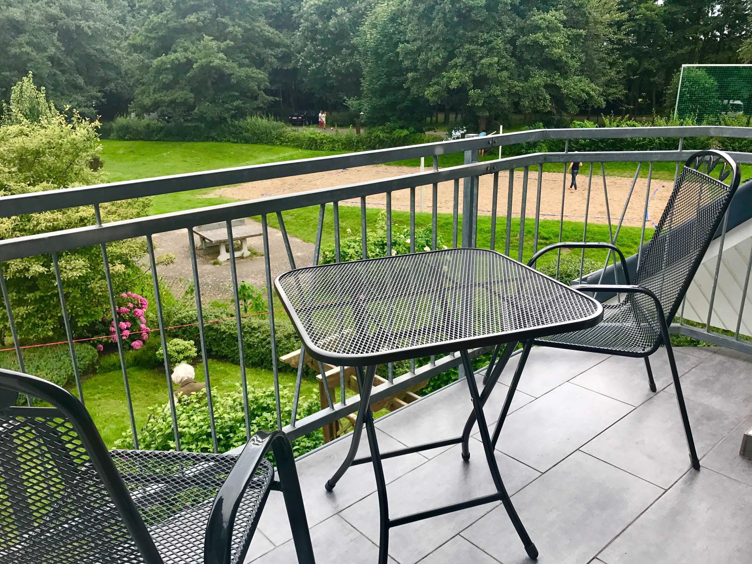 Un balcon avec deux chaises en métal et une petite table, donnant sur un jardin avec un court de tennis et des arbres.