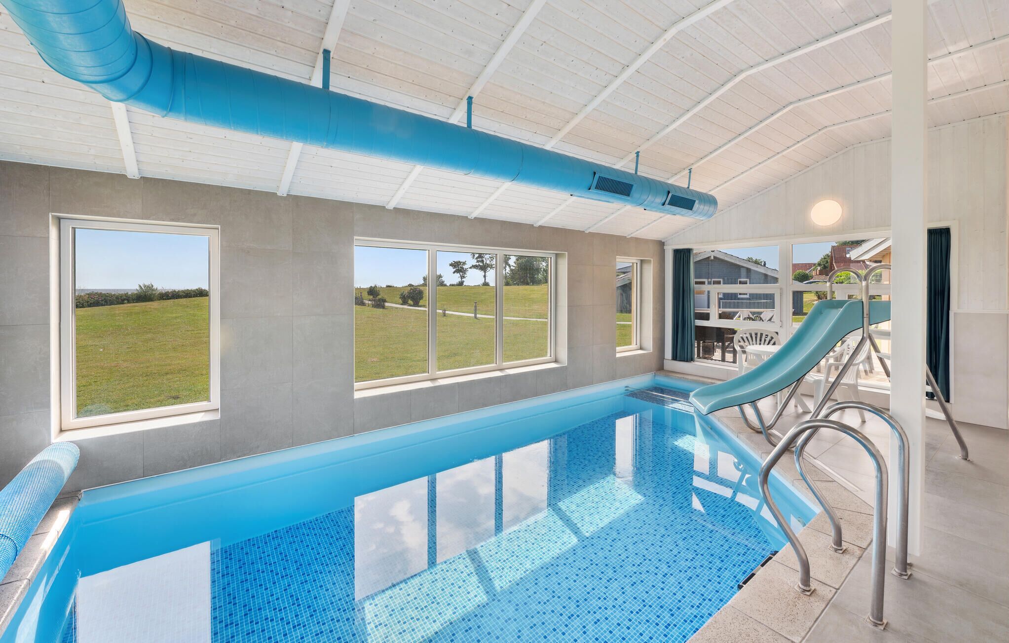A modern indoor swimming pool with clear glass walls, a blue pool edge, and a view of a grassy field.