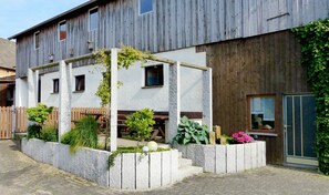Un bâtiment à deux étages doté d’une façade en bois, d’un petit jardin avec des plantes, d’une structure en béton et d’une clôture en bois.