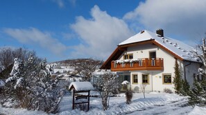 Une maison recouverte de neige, dotée d’un balcon, entourée d’arbres et surplombant un ciel dégagé.