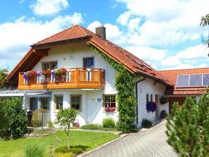 Une maison à deux étages avec un toit recouvert de tuiles rouges, un balcon doté de balustrades en bois et un jardin à l’avant.
