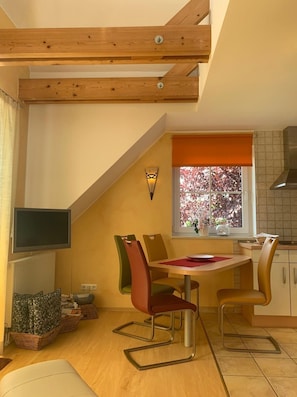 A dining area with a table and chairs, a television mounted on the wall, and a window with a view of trees. - Holiday house Wölkchen - holiday house (Zinnowitz)