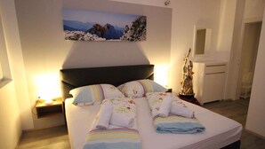 Una cama bien tendida con toallas, una mesita de noche con una lámpara, un cuadro de montañas y un espejo en la pared.