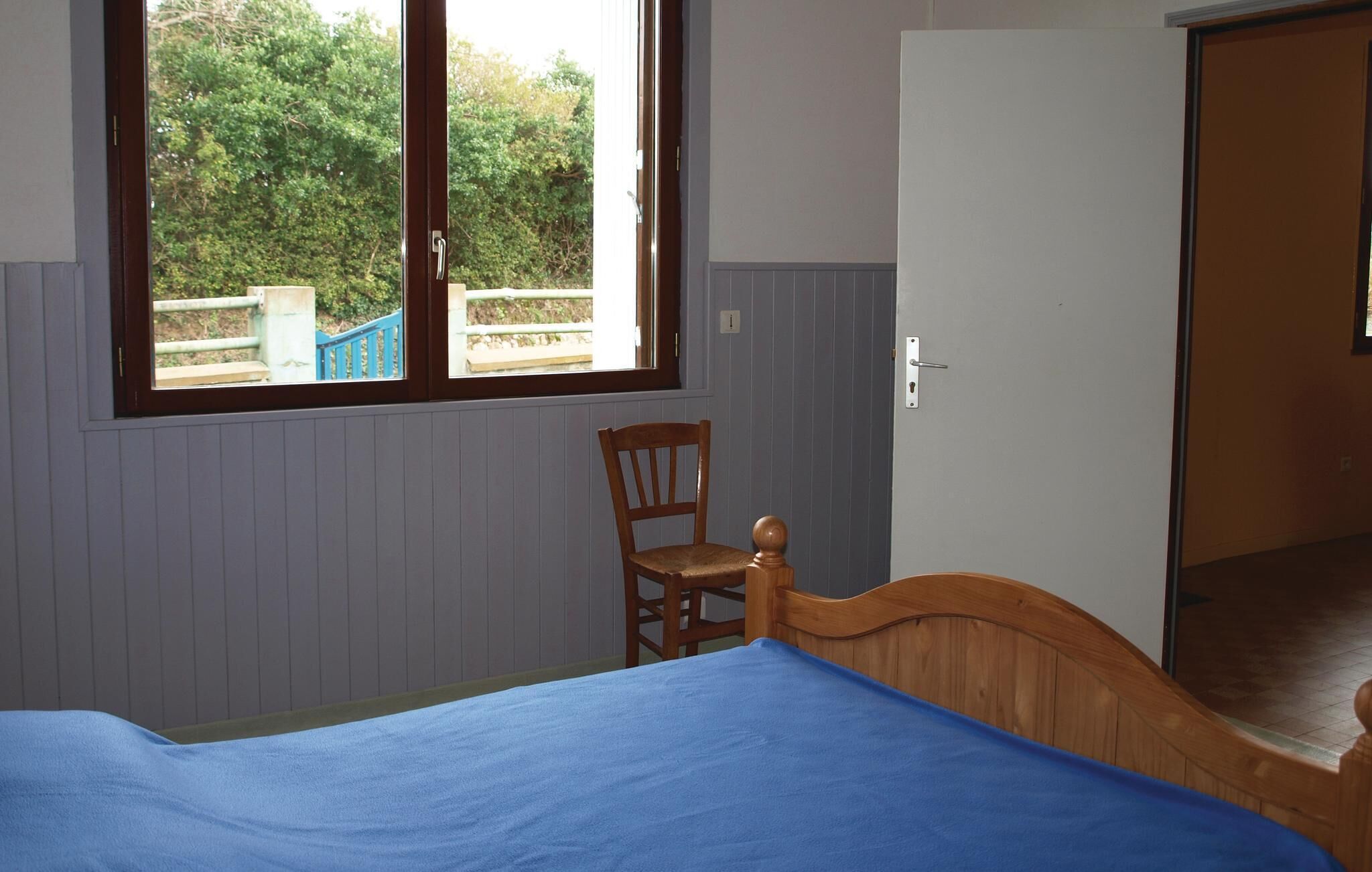 Ein Schlafzimmer mit einem Einzelbett, einem Holzsessel, einem Fenster mit Blick auf Grünflächen und einer Tür.