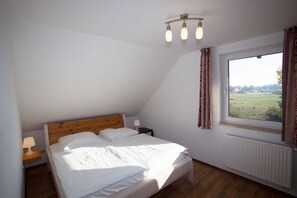 Et soveværelse med træsenggavl, en enkeltseng, et natbord med lampe, et vindue med gardiner og udsigt til et grønt landskab.