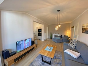 A modern living room with a flat-screen TV, a wooden coffee table, a grey sofa, and a kitchen in the background.