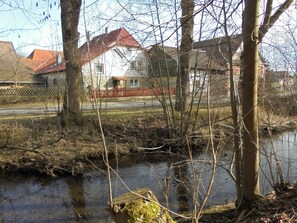 Ein Fluss mit einer Holzbrücke, ein weißes Haus mit rotem Dach und ein Zaun im Hintergrund.