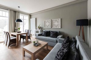 Un salon moderne avec un canapé gris, une table basse en bois, un coin repas avec une table et des chaises, et des œuvres d’art décoratives accrochées au mur.