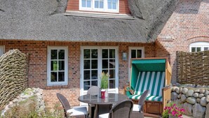 Une maison en briques avec un toit de chaume, une table ronde entourée de chaises et un espace extérieur aménagé avec des sièges à rayures vertes.