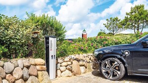 Un VUS noir est garé à côté d’une borne de recharge, dans un espace aménagé avec un mur de pierres, de la verdure et un phare en arrière-plan.