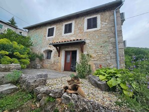 Une maison en pierre avec une porte en bois, un petit porche et un jardin agrémenté de diverses plantes et bordé d’un mur en pierre.
