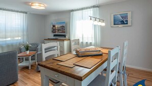 A modern living room with a wooden dining table, white chairs, a grey sofa, and a TV mounted on the wall. - Blue hour, Germany (Heringsdorf)