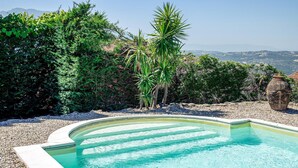 A swimming pool with a curved edge, surrounded by gravel, and bordered by dense greenery and a view of distant hills. - Nice home in Contursi Terme (SA) with sauna (Contursi Terme (SA))