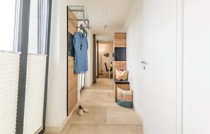 A narrow hallway with a wooden wardrobe, a hanging rack with clothes, a bench with a bag, and a door leading to another room.