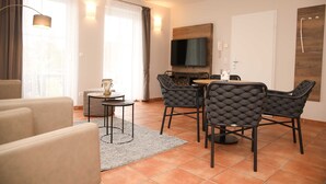 A modern living room with a sofa, a dining table with chairs, a TV mounted on the wall, and a window with curtains. - Apartment with Jacuzzi (5) - Apartment near the beach (Nienhagen)