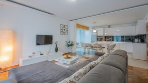 Un saló modern amb un sofà gris, una taula de centre i una televisió. Hi ha una zona de dinar amb caires i taula, i una cuina amb armaris blancs i electrodomèstics.