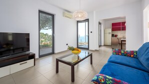 Un salon moderne avec un téléviseur, une table basse en verre sur laquelle sont disposés des fruits, un canapé bleu et une vue sur la mer grâce à de grandes fenêtres.