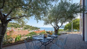 Un espace de restauration en plein air avec une table dressée pour quatre personnes, offrant une vue sur la côte, avec des maisons et des bateaux visibles au loin.