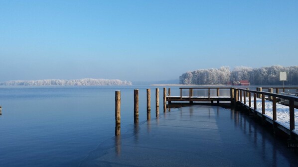 Un lago congelado con un muelle de madera que se adentra en el agua, rodeado de árboles cubiertos de nieve y un cielo despejado.