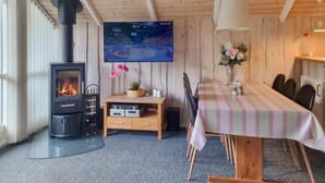 Een gezellige kamer met een houtkachel, een aan de muur bevestigde televisie, een eettafel met stoelen en een klein kastje.