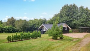 Een klein, gelijkvloers huis met een donker dak en witte accenten, omgeven door een verzorgd gazon en een rij jonge bomen.