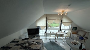 A bedroom with a bed, a TV, a desk, a chair, a sofa, and a view of the outdoors. - 1-room apartment 1, 30 m² - Graf Luckner Hüs (Scharbeutz)