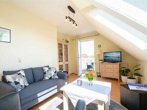 A modern living room with a grey sofa, a white coffee table, and a TV on a wooden cabinet. There is a bookshelf and a window with a view of the outside. - Meadow ground 18 Whg. WiGr-18 - meadow ground 18 (Kühlungsborn)