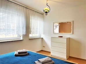 Een slaapkamer met een bed, een dressoir, een spiegel en een raam met dunne gordijnen.