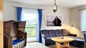 Un salon moderne avec un canapé, une table basse en bois, un fauteuil en osier et une vue sur l’extérieur grâce à une grande fenêtre.