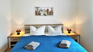 Un lit bien fait, recouvert d’une couverture bleue, avec deux oreillers et deux serviettes pliées. On y trouve deux lampes de chevet, une tête de lit en bois et un tableau représentant un paysage urbain accroché au mur.