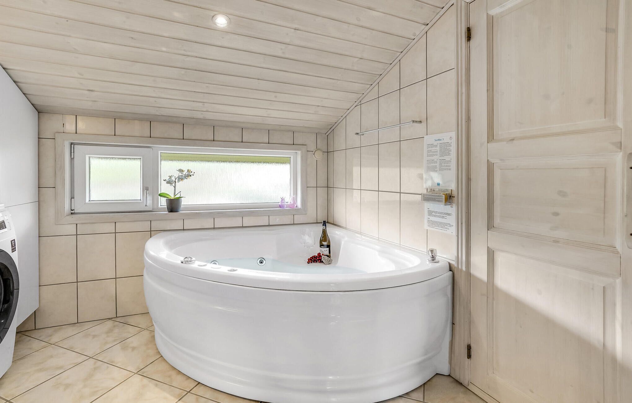 A bathroom with a large white bathtub, a window with plants, and a door with a sign.