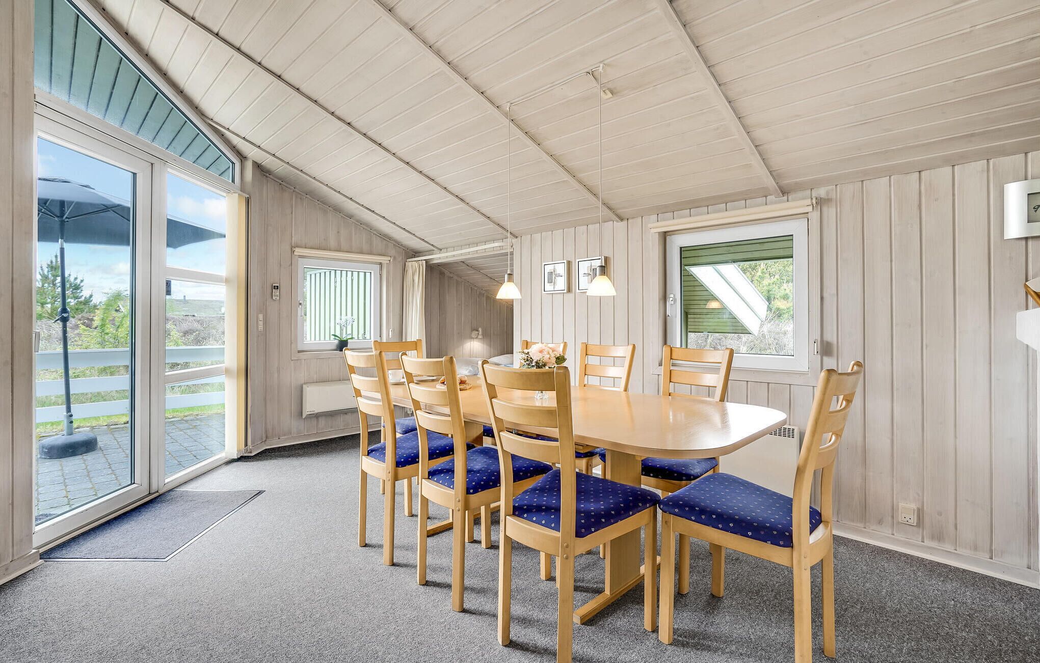Un espace repas avec une table ronde et six chaises, des murs en bois et de grandes fenêtres.