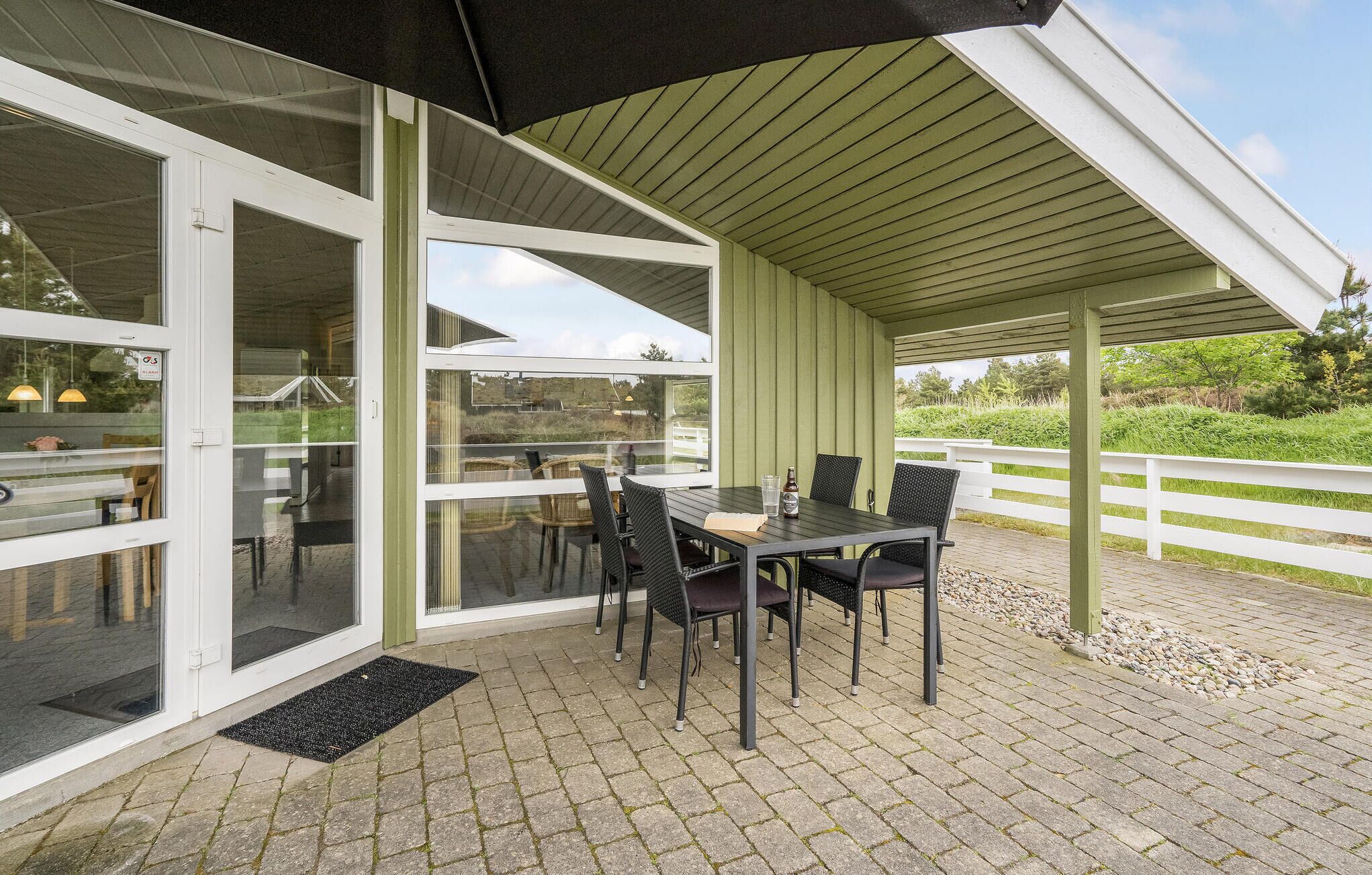 An outdoor patio with a table and chairs, a covered area with a roof, and a white fence in the background.